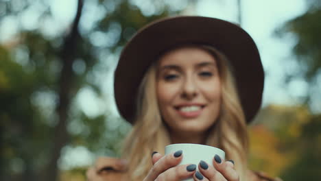 Bottom-view-of-Caucasian-woman-in-a-hat-holding-a-white-cup-of-coffee-and-looking-at-the-camera-in-the-park-in-autumn