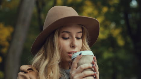 Close-up-view-of-Caucasian-young-blonde-woman-in-a-hat-drinking-hot-coffee-sitting-on-a-bench-in-the-park-in-autumn