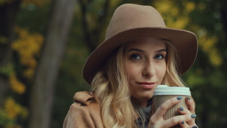 Close-up-view-of-Caucasian-young-blonde-woman-in-a-hat-drinking-hot-coffee-sitting-on-a-bench-while-looking-at-the-camera-in-the-park-in-autumn