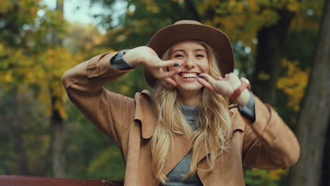 Caucasian-young-blonde-woman-in-a-hat-and-coat-laughing-while-posing-to-the-camera-and-doing-funny-muzzles-and-gestures-in-the-park-in-autumn