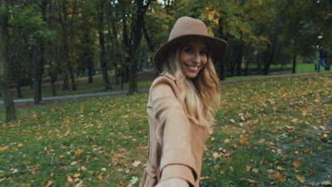 Rear-view-of-Caucasian-young-blonde-woman-in-a-hat-and-coat-walking-in-the-park-and-leading-the-camera-with-her,-then-she-turns-and-smiles