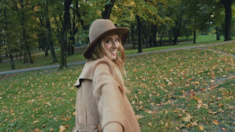 Rear-view-of-Caucasian-young-blonde-woman-in-a-hat-and-coat-walking-in-the-park-and-leading-the-camera-with-her,-then-she-turns-and-smiles
