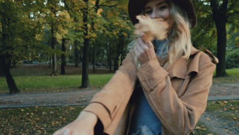 Rear-view-of-Caucasian-young-blonde-woman-in-a-hat-and-coat-walking-in-the-park-and-leading-the-camera-with-her,-then-she-turns-and-smiles-while-holding-a-yellow-leaf