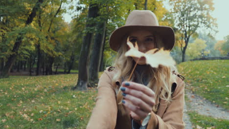 Rear-view-of-Caucasian-young-blonde-woman-in-a-hat-and-coat-walking-in-the-park-and-leading-the-camera-with-her,-then-she-turns-and-smiles-while-holding-a-yellow-leaf