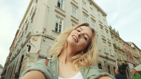 Close-up-view-of-caucasian-blonde-young-woman-smiling-and-posing-to-the-camera-in-the-street-while-holding-it