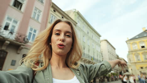 Close-up-view-of-caucasian-blonde-young-woman-speaking-and-waving-to-the-camera-in-the-street-while-holding-it