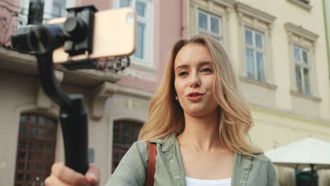 Close-up-view-of-caucasian-blonde-young-woman-recording-a-video-on-the-smartphone-while-holding-it-with-a-selfie-stick-in-the-street