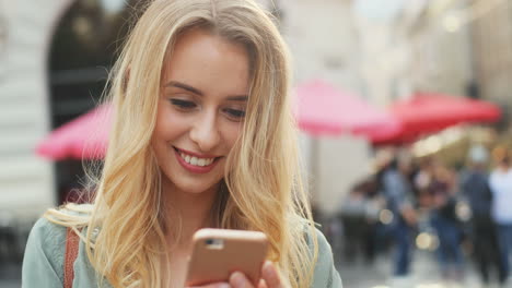 Close-up-view-of-Caucasian-blonde-woman-texting-on-the-smartphone-and-smiling-in-the-street