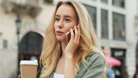 Close-up-view-of-Caucasian-blonde-woman-talking-on-the-smartphone-while-walking-down-the-street-and-drinking-coffee-to-go