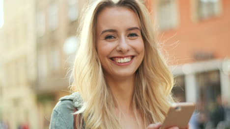 Close-up-view-of-young-blonde-woman-texting-on-her-smartphone-and-looking-at-the-camera-in-the-street
