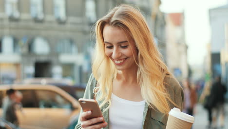 Close-up-view-of-Caucasian-blonde-woman-texting-on-the-smartphone-while-walking-down-the-street-and-holding-coffee-to-go