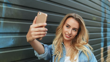 Young-Caucasian-blonde-woman-in-jeans-jacket-taking-a-selfie-on-the-smartphone-in-the-street