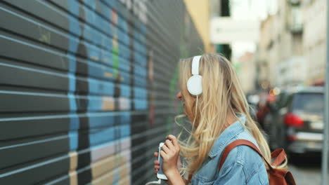 Caucasian-blonde-young-woman-listening-to-music-with-headphones-and-dancing-in-the-street