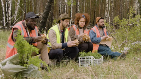 Group-of-multiethnic-ecologist-activists-eating-and-talking-in-a-break-sitting-on-a-trunk-in-the-forest