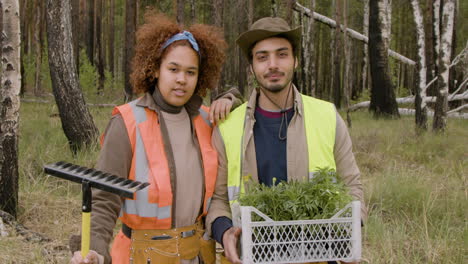 African-american-woman-activist-and-caucasian-male-coworker-smiling-at-the-camera-while-they-holding-a-rake-and-small-trees