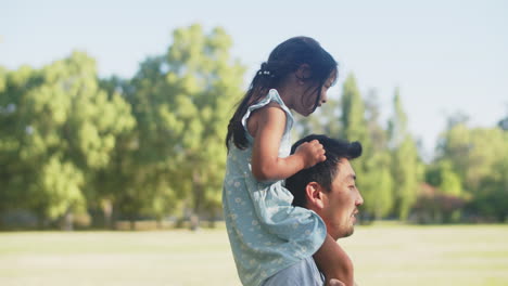 Side-view-of-happy-Asian-girl-riding-father-piggyback-in-park