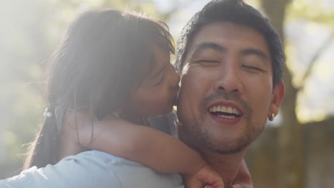 Close-up-of-cute-little-Asian-girl-on-back-of-her-father,-smiling-and-kissing-him