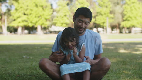 Happy-young-Asian-father-tickling-his-little-daughter-outdoors