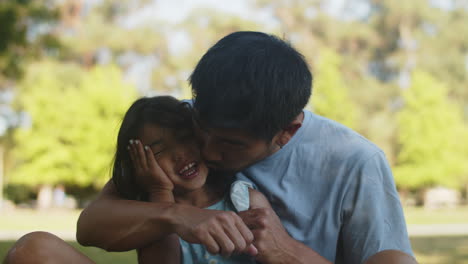 Happy-Asian-father-sitting-with-little-daughter-on-the-grass-and-kissing-her