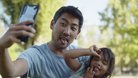 Hombre-Asiático-Feliz-Tomando-Selfie-Con-Su-Hija-Con-Teléfono-Inteligente-En-El-Parque