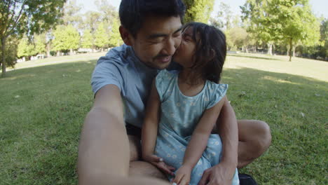 Happy-father-sitting-on-grass-with-daughter-and-taking-video-while-the-girl-kisses-him
