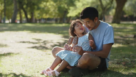 Cute-little-daughter-sitting-with-father-on-grass-and-eating-chocolate-ice-creams