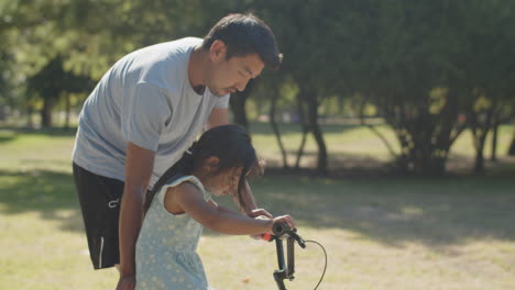 Padre-Joven-Feliz-Enseñando-A-Su-Hija-A-Andar-En-Bicicleta-En-El-Parque