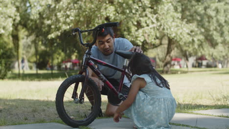 Curious-little-daughter-spinning-pedal-of-her-bike-in-park-with-her-father