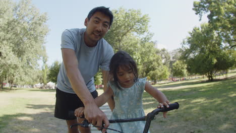Niña-Asiática-Feliz-Aprendiendo-A-Andar-En-Bicicleta-Con-Su-Padre-En-El-Parque