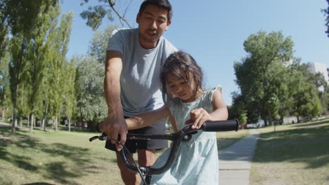 Little-Asian-girl-riding-bike-with-her-father-in-park