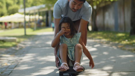 Young-father-giving-a-ride-on-skateboard-to-his-little-daughter