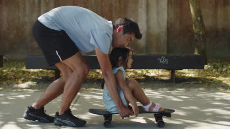 Happy-young-father-moving-skateboard-with-little-daughter