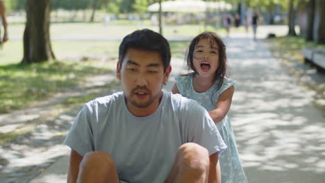 Glückliches-Asiatisches-Kleines-Mädchen,-Das-Seinen-Vater-Auf-Dem-Skateboard-Sitzt