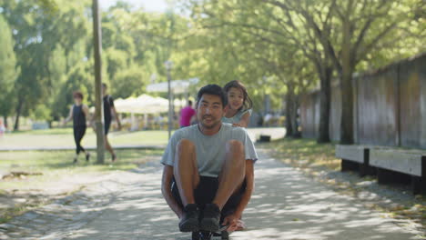 Happy-Asian-man-sitting-on-skateboard,-daughter-pushing-him
