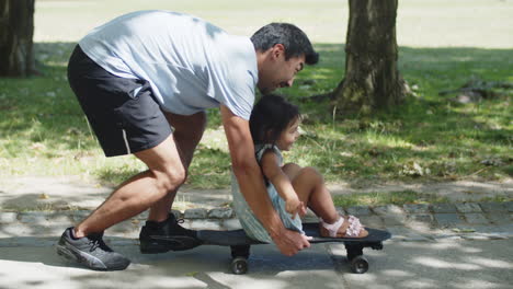 Side-view-of-happy-Asian-man-giving-his-daughter-ride-on-skateboard-in-park