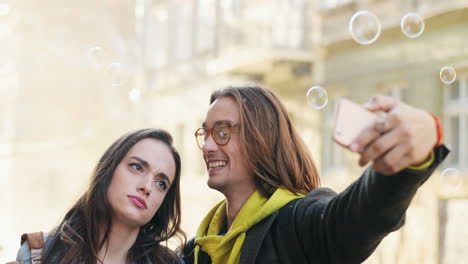 Close-up-view-of-young-Caucasian-couple-of-friends-taking-selfies-while-transparent-balls-of-soap-falling