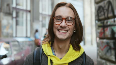 Close-up-view-of-young-Caucasian-man-with-long-hair-and-wearing-glasses-smiling-to-the-camera-in-the-street