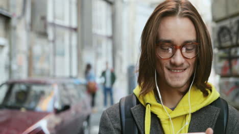 Close-up-view-of-Caucasian-young-man-in-stylish-look-and-headphones-smiling-and-typing-on-the-smartphone-in-the-street
