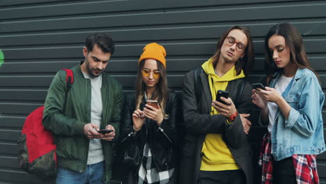 Group-of-caucasian-friends-leaning-on-the-wall-while-talking-and-typing-on-their-smartphones-in-the-street
