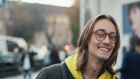 Close-up-view-of-good-looking-young-caucasian-man-in-glasses-looking-aside-while-standing-outdoor-in-the-city,-then-turns-his-head-and-smiling-at-the-camera