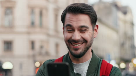 Close-up-view-of-young-Caucasian-man-with-backpack-texting-on-his-smartphone-cheerfully-in-the-street