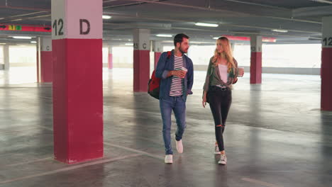 Young-Caucasian-and-stylish-man-and-woman-walking-in-a-empty-parking-and-talking-while-drinking-coffee