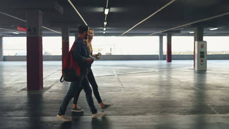 Side-view-of-young-Caucasian-and-stylish-man-and-woman-walking-in-a-empty-parking-and-talking-while-drinking-coffee