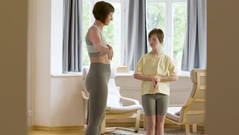 Brunette-mother-in-sporstwear-teaching-yoga-to-her-daughter-with-down-syndrome-at-home