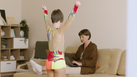 Young-girl-in-gymnastics-dress-practicing-in-front-of-her-mother,-who-is-using-a-laptop