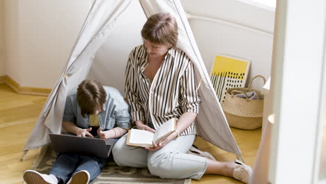 Mother-reading-book-at-home-next-to-her-daughter-using-laptop