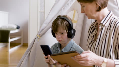 Teenager-with-down-syndrome-using-headphones-and-mobile-phone,-and-talking-with-her-mother,-who-is-using-tablet