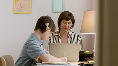 Mature-woman-in-striped-shirt-working-with-laptop-and-her-daughter-is-doing-her-homework