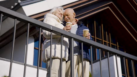 Seniorenpaar-Umarmt-Sich,-Während-Es-Kaffee-Hält,-Um-An-Einem-Wintertag-Auf-Eine-Terrasse-Im-Park-Zu-Gehen