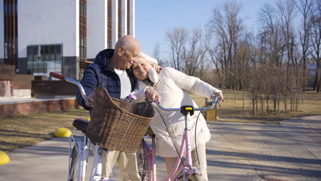 Senior-couple-holding-bikes-while-looking-to-each-other,-hugging-and-smiling-in-the-park-on-a-winter-day